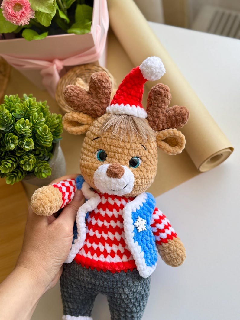 May include: A crocheted reindeer doll wearing a red and white striped sweater, a blue vest with a snowflake, and gray pants. The reindeer has brown antlers, a red Santa hat with a white pom-pom, and blue eyes. A festive holiday decoration.