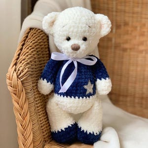 May include: A white crocheted teddy bear wearing a blue sweater with a silver star. The bear is sitting on a wicker chair with a white blanket.