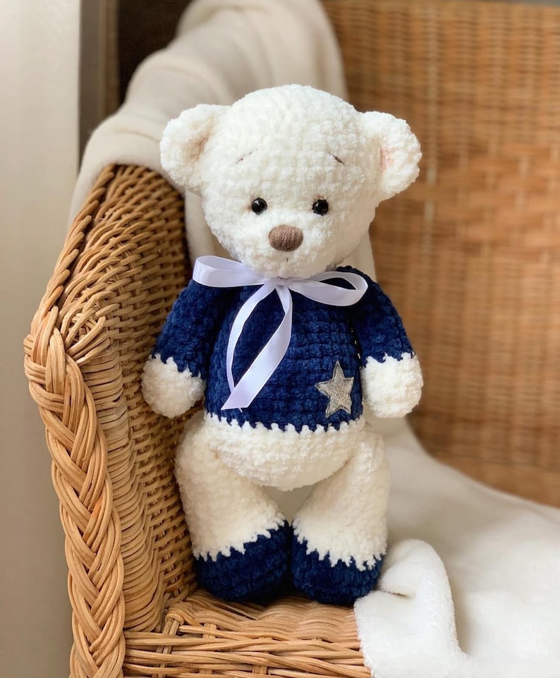 May include: A white crocheted teddy bear wearing a blue sweater with a silver star. The bear is sitting on a wicker chair with a white blanket.