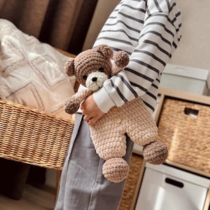 May include: A child holding a brown and beige crocheted teddy bear wearing a matching outfit. The teddy bear has a white face and black eyes.