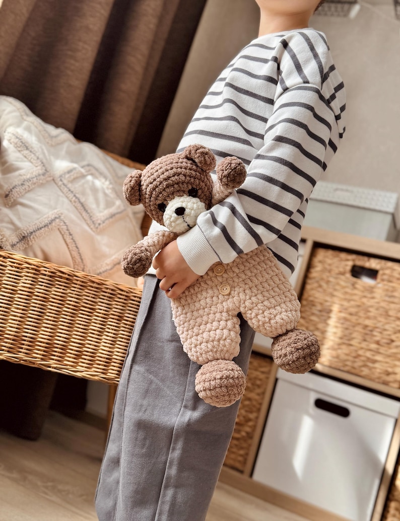 May include: A child holding a brown and beige crocheted teddy bear wearing a matching outfit. The teddy bear has a white face and black eyes.