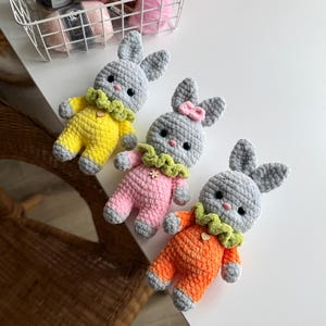 May include: Three crocheted bunny plush toys. The bunnies are gray with different colored outfits. One bunny is wearing a yellow outfit, one is wearing a pink outfit, and one is wearing an orange outfit.