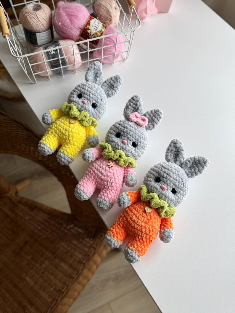 May include: Three crocheted bunny plush toys. The bunnies are gray with different colored outfits. One bunny is wearing a yellow outfit, one is wearing a pink outfit, and one is wearing an orange outfit.