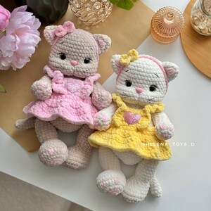 Pode incluir: Duas bonecas de gato de croch&ecirc; feitas &agrave; m&atilde;o. Uma &eacute; bege com um vestido e la&ccedil;o rosa, a outra &eacute; branca com um vestido e la&ccedil;o amarelo. Ambas as bonecas t&ecirc;m enfeites em forma de cora&ccedil;&atilde;o em seus vestidos e est&atilde;o em uma superf&iacute;cie branca.