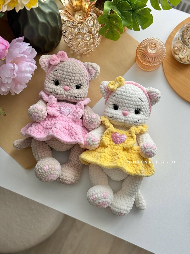 Pode incluir: Duas bonecas de gato de croch&ecirc; feitas &agrave; m&atilde;o. Uma &eacute; bege com um vestido e la&ccedil;o rosa, a outra &eacute; branca com um vestido e la&ccedil;o amarelo. Ambas as bonecas t&ecirc;m enfeites em forma de cora&ccedil;&atilde;o em seus vestidos e est&atilde;o em uma superf&iacute;cie branca.