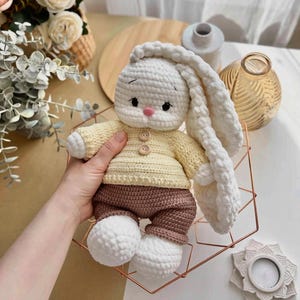May include: A handmade crochet bunny doll with long, floppy ears. The bunny wears a yellow sweater and brown pants. The doll has a pink nose and button details. The bunny is sitting in a copper-colored geometric wire basket.