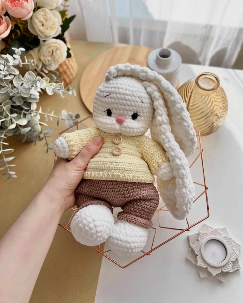 May include: A handmade crochet bunny doll with long, floppy ears. The bunny wears a yellow sweater and brown pants. The doll has a pink nose and button details. The bunny is sitting in a copper-colored geometric wire basket.