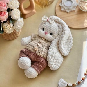 May include: A handmade crochet bunny doll with long, floppy ears and a small bow. The bunny wears a tan sweater with wooden buttons and pink pants. The doll is white, tan, and pink, and is a soft toy.