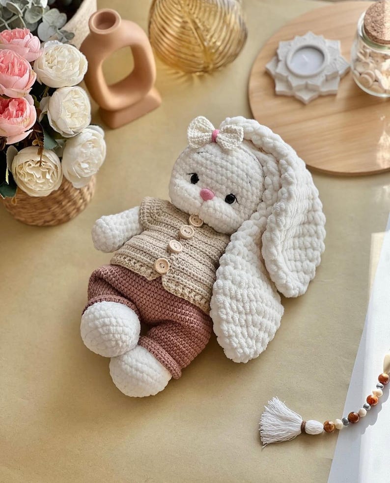 May include: A handmade crochet bunny doll with long, floppy ears and a small bow. The bunny wears a tan sweater with wooden buttons and pink pants. The doll is white, tan, and pink, and is a soft toy.