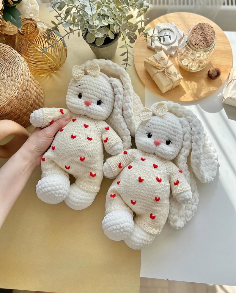 Puede incluir: Dos mu&ntilde;ecos de conejito de ganchillo hechos a mano, de color crema, con largas orejas ca&iacute;das y peque&ntilde;os acentos de coraz&oacute;n rojo. Cada conejito tiene un peque&ntilde;o lazo en la cabeza y una nariz rosa. Los conejitos son suaves y tiernos.