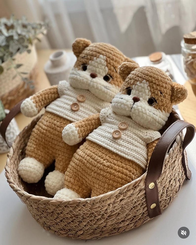 Pu&ograve; includere: Due cani all'uncinetto fatti a mano in un cesto di vimini. I cani sono marrone chiaro e bianchi, con maglioni color crema con bottoni di legno. Il cesto ha manici in pelle marrone.