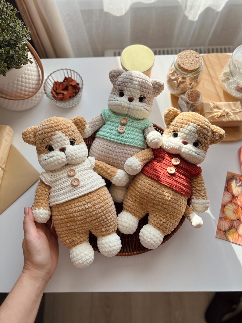 Pu&ograve; includere: Tre bambole a forma di gatto all'uncinetto fatte a mano in un cestino intrecciato. Ogni gatto ha una combinazione di colori marrone e bianco, con maglioni di diversi colori: bianco, verde chiaro e rosso. Le bambole hanno dettagli di bottoni e sono esposte su una superficie bianca.