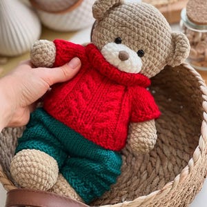 May include: A handmade crochet teddy bear wearing a red sweater and green pants. The bear has a tan body, a white muzzle, and black eyes. The bear is sitting in a woven basket.
