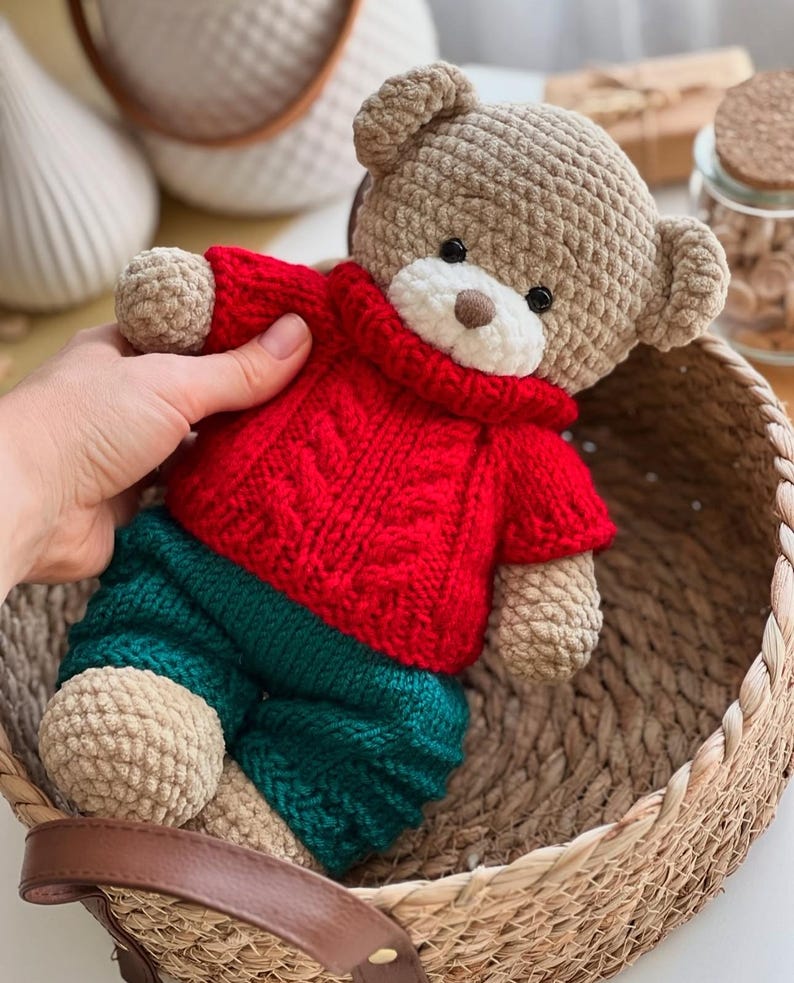 May include: A handmade crochet teddy bear wearing a red sweater and green pants. The bear has a tan body, a white muzzle, and black eyes. The bear is sitting in a woven basket.