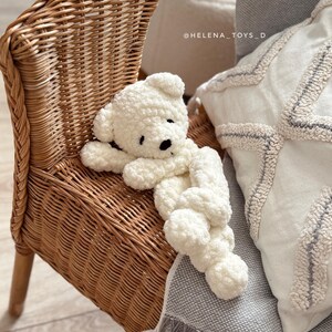 Pode incluir: Um urso de pel&uacute;cia branco, com olhos e nariz pretos, deitado em uma cadeira de vime. O urso &eacute; feito de um material macio e texturizado. A cadeira &eacute; de cor castanho claro e tem um design tecido. Uma almofada branca e um cobertor cinza est&atilde;o no fundo.