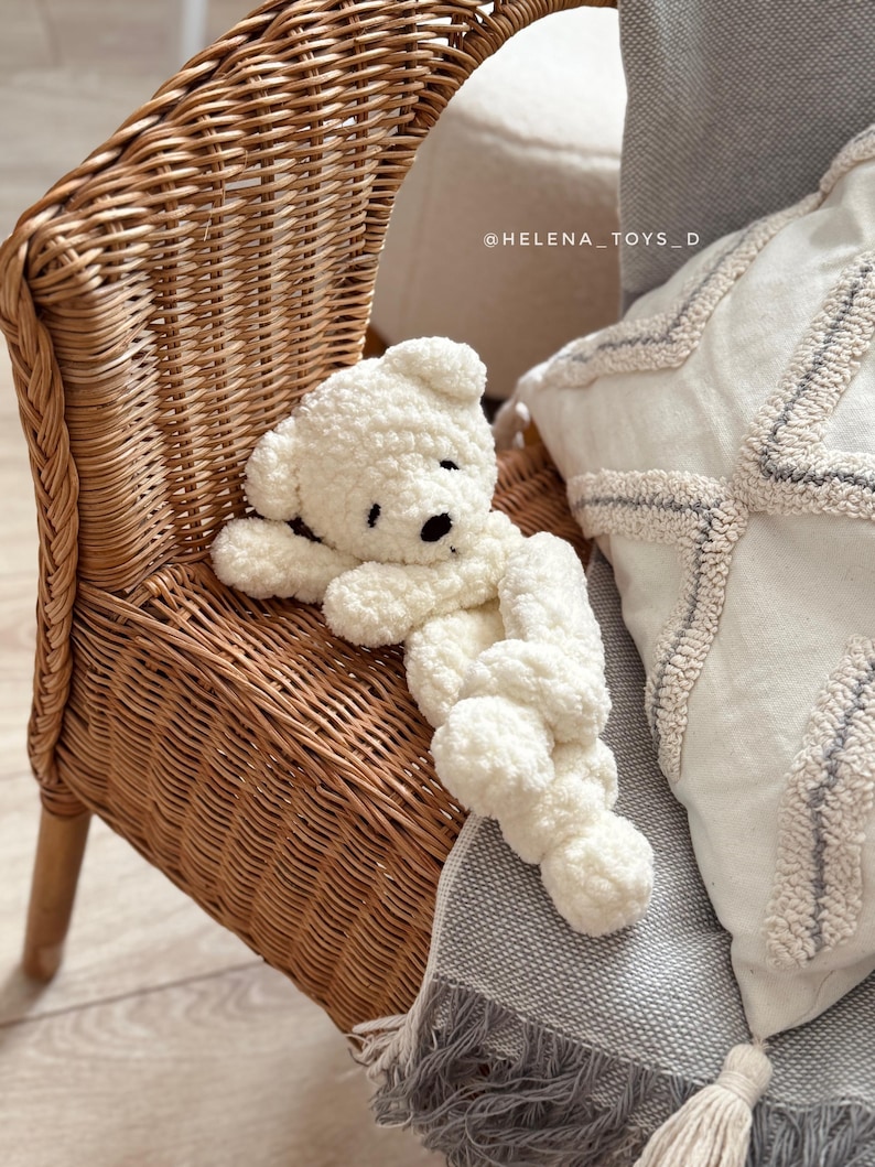 Pode incluir: Um urso de pel&uacute;cia branco, com olhos e nariz pretos, deitado em uma cadeira de vime. O urso &eacute; feito de um material macio e texturizado. A cadeira &eacute; de cor castanho claro e tem um design tecido. Uma almofada branca e um cobertor cinza est&atilde;o no fundo.