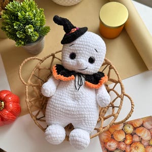 May include: A handmade white crochet ghost doll wearing a black witch's hat with orange and purple details. The ghost has a black collar with orange trim and sits in a woven basket. An orange pumpkin and a green plant are also visible.