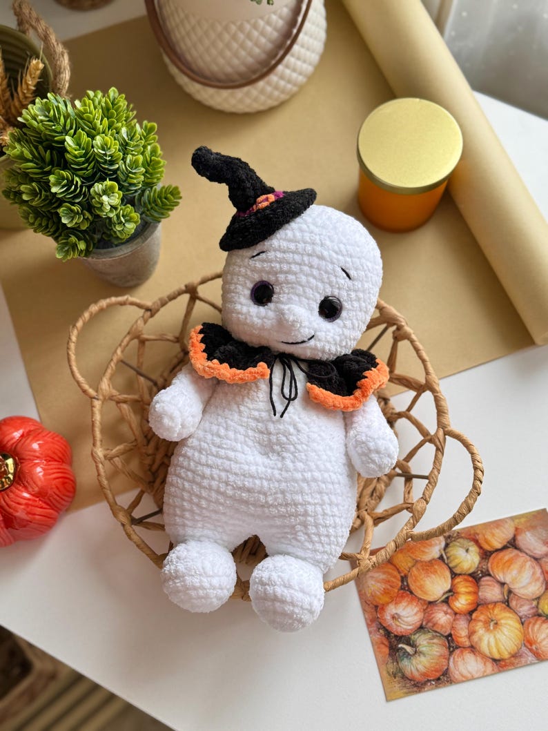 May include: A handmade white crochet ghost doll wearing a black witch's hat with orange and purple details. The ghost has a black collar with orange trim and sits in a woven basket. An orange pumpkin and a green plant are also visible.