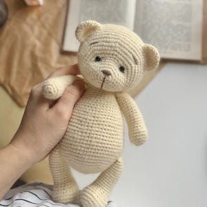 May include: A cream-colored, crocheted teddy bear with black eyes and a brown nose. The handmade bear has a classic design with rounded features and is being held by a person. The bear is a soft toy.