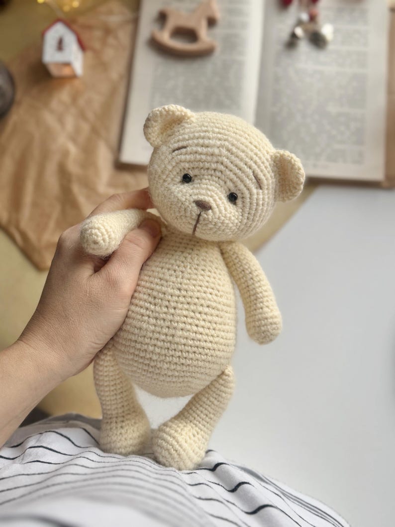 May include: A cream-colored, crocheted teddy bear with black eyes and a brown nose. The handmade bear has a classic design with rounded features and is being held by a person. The bear is a soft toy.