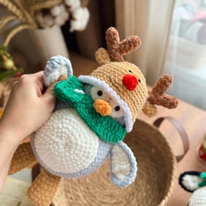 May include: A handmade crochet reindeer toy with a red nose, brown antlers, and a tan hat. The reindeer is wearing a green scarf and has a white and blue body. The toy is soft and plush, perfect for holiday decor or a child's gift.