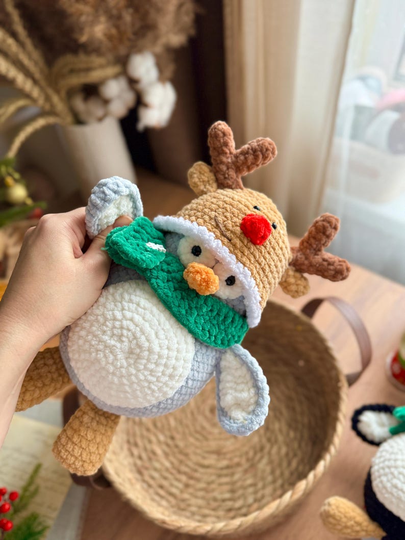 May include: A handmade crochet reindeer toy with a red nose, brown antlers, and a tan hat. The reindeer is wearing a green scarf and has a white and blue body. The toy is soft and plush, perfect for holiday decor or a child's gift.