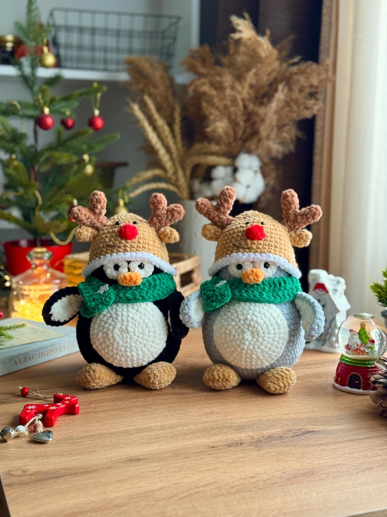 May include: Two handmade crochet penguin figures, one black and white, the other gray and white. Both wear reindeer hats with brown antlers and red noses, and green scarves. They are Christmas-themed decorations.