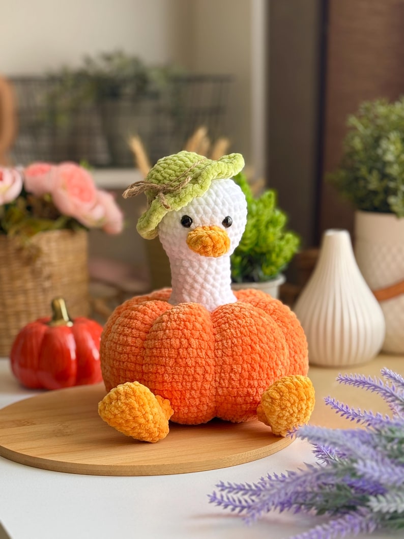 May include: A handmade crochet duck wearing a green hat, with an orange pumpkin body and feet. The duck has a white head, orange beak, and black eyes. The duck is sitting on a wooden surface.