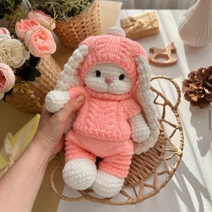 May include: A handmade pink and white crochet bunny doll wearing a matching hat and outfit. The bunny has long, floppy ears and is sitting in a woven basket. The doll is soft and cuddly, perfect for a child's toy or nursery decor.