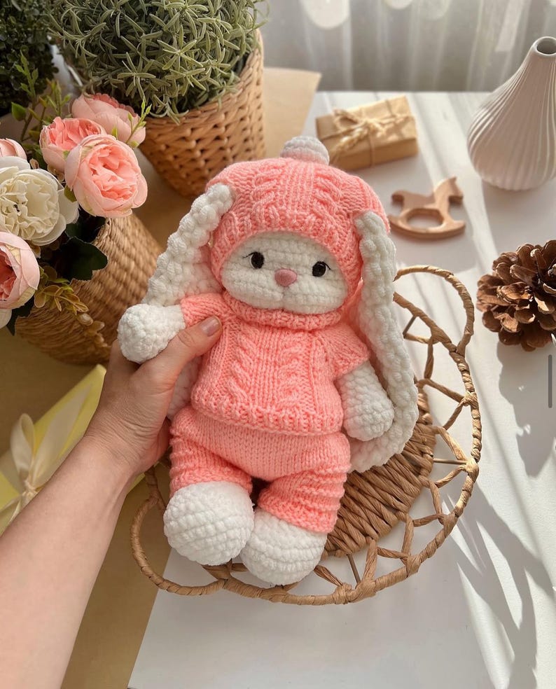 May include: A handmade pink and white crochet bunny doll wearing a matching hat and outfit. The bunny has long, floppy ears and is sitting in a woven basket. The doll is soft and cuddly, perfect for a child's toy or nursery decor.