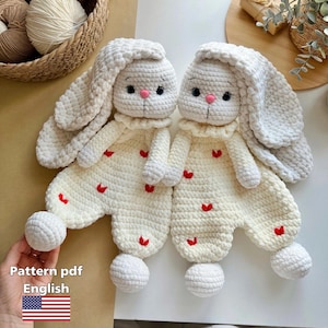 May include: Two handmade, white crochet bunny dolls with long ears and pink noses. The bunnies wear cream-colored outfits adorned with red hearts. The image includes text that says "Pattern pdf English".