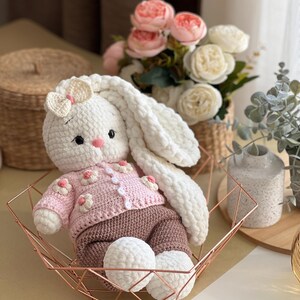 May include: A handmade white crochet bunny wearing a pink sweater with flower accents and brown pants. The bunny has long, floppy ears and is seated in a rose gold geometric basket. The bunny's feet are white pom-poms.