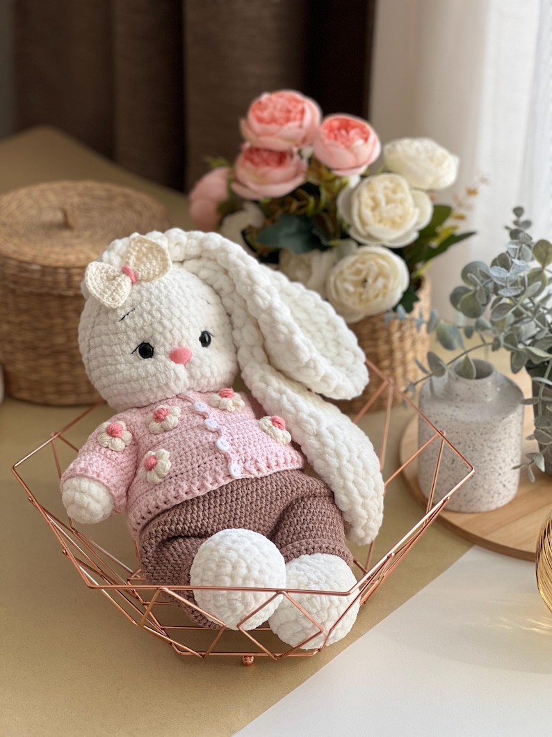 May include: A handmade white crochet bunny wearing a pink sweater with flower accents and brown pants. The bunny has long, floppy ears and is seated in a rose gold geometric basket. The bunny's feet are white pom-poms.