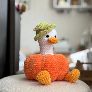 May include: A handmade crochet duck wearing a green leaf hat and an orange pumpkin body. The duck has a white head, orange beak, and small black eyes. The duck is sitting on a white, textured surface.