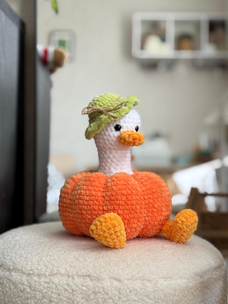 May include: A handmade crochet duck wearing a green leaf hat and an orange pumpkin body. The duck has a white head, orange beak, and small black eyes. The duck is sitting on a white, textured surface.