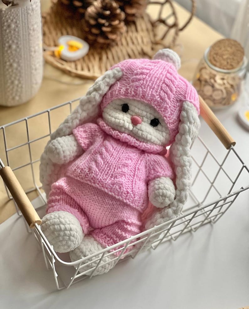 May include: A handmade, pink and white knitted bunny doll wearing a matching hat and outfit. The bunny is in a white wire basket with wooden handles. The doll has long white ears and a pink nose.