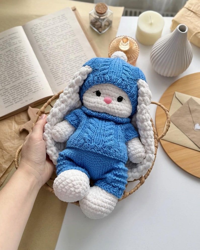 May include: A handmade crochet bunny doll in a blue sweater and pants set, with a matching hat. The bunny has white ears and feet, a pink nose, and black eyes. The doll is in a woven basket.