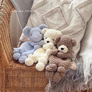 Pode incluir: Tr&ecirc;s bichos de pel&uacute;cia de croch&ecirc; feitos &agrave; m&atilde;o: um rato azul, um urso creme e um urso marrom. Eles est&atilde;o descansando em uma cadeira de vime com uma almofada decorativa e um cobertor com franjas. Os brinquedos s&atilde;o macios e fofinhos.