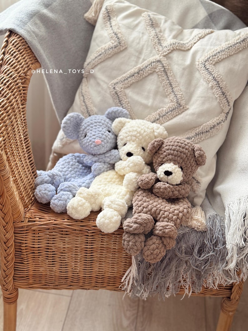 Pode incluir: Tr&ecirc;s bichos de pel&uacute;cia de croch&ecirc; feitos &agrave; m&atilde;o: um rato azul, um urso creme e um urso marrom. Eles est&atilde;o descansando em uma cadeira de vime com uma almofada decorativa e um cobertor com franjas. Os brinquedos s&atilde;o macios e fofinhos.