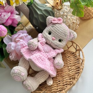 Pode incluir: Boneca de gato de croch&ecirc; feita &agrave; m&atilde;o, com um vestido rosa com um desenho de cora&ccedil;&atilde;o. A boneca tem um corpo cinza claro, detalhes rosa e um la&ccedil;o a condizer. Descansa numa cesta de vime, rodeada de flores e vegeta&ccedil;&atilde;o.