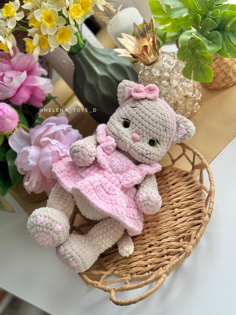 Pode incluir: Boneca de gato de croch&ecirc; feita &agrave; m&atilde;o, com um vestido rosa com um desenho de cora&ccedil;&atilde;o. A boneca tem um corpo cinza claro, detalhes rosa e um la&ccedil;o a condizer. Descansa numa cesta de vime, rodeada de flores e vegeta&ccedil;&atilde;o.