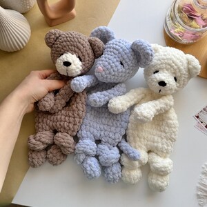 Pode incluir: Tr&ecirc;s ursos de pel&uacute;cia de croch&ecirc; feitos &agrave; m&atilde;o em marrom, azul e branco. Cada urso tem um nariz preto e um focinho branco. Os ursos est&atilde;o em uma superf&iacute;cie branca. Os ursos s&atilde;o macios e fofinhos.