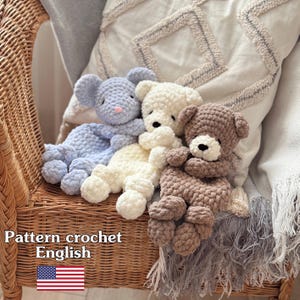 May include: Three handmade crochet teddy bears in light blue, cream, and brown. The bears are sitting on a wicker chair with a decorative pillow in the background. The text "Pattern crochet English" is visible in the image.