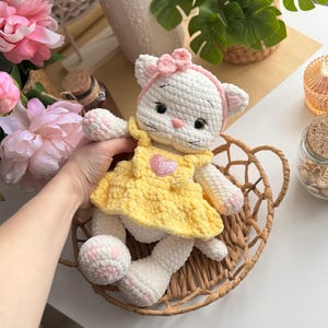 Pode incluir: Boneca de gato de croch&ecirc; branco feita &agrave; m&atilde;o com uma faixa de la&ccedil;o rosa e um vestido amarelo. A boneca tem detalhes rosa nas patas e um cora&ccedil;&atilde;o no vestido. Est&aacute; sentada em uma cesta de vime.