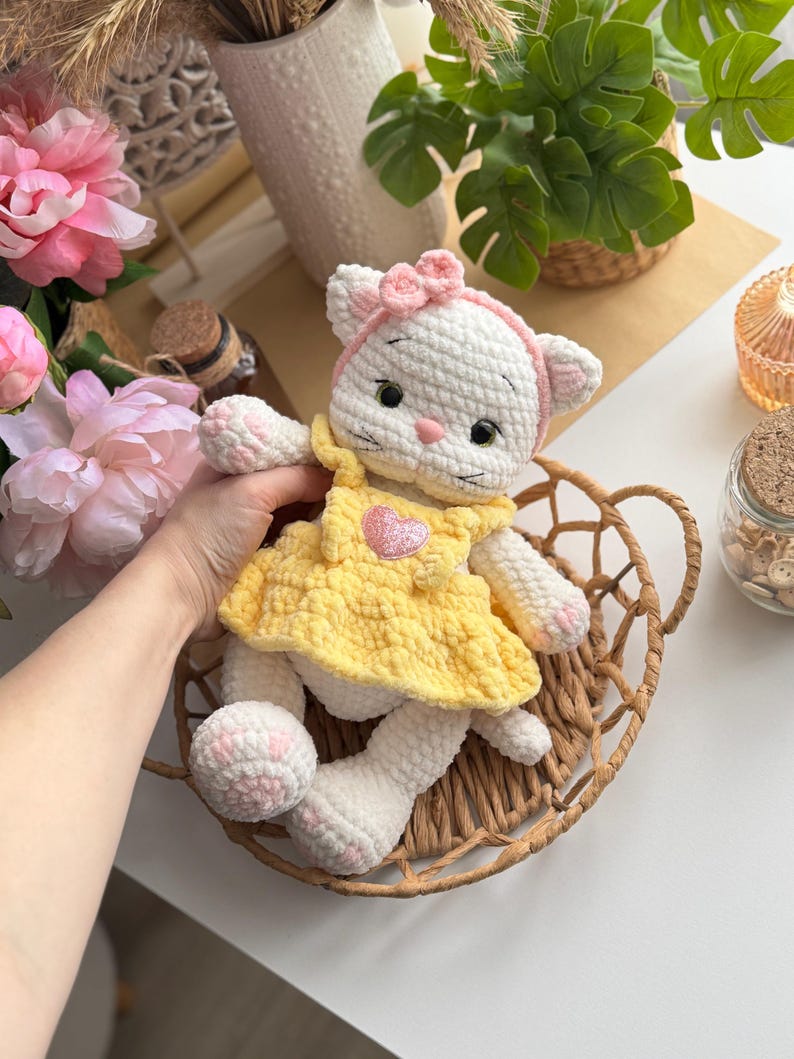 Pode incluir: Boneca de gato de croch&ecirc; branco feita &agrave; m&atilde;o com uma faixa de la&ccedil;o rosa e um vestido amarelo. A boneca tem detalhes rosa nas patas e um cora&ccedil;&atilde;o no vestido. Est&aacute; sentada em uma cesta de vime.