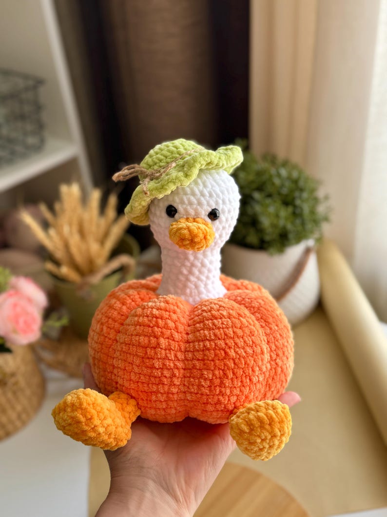 May include: A handmade crochet goose doll with a white head, orange beak, and a green leaf hat. The goose is seated in an orange pumpkin body with orange feet. A whimsical, soft toy for children or home decor.