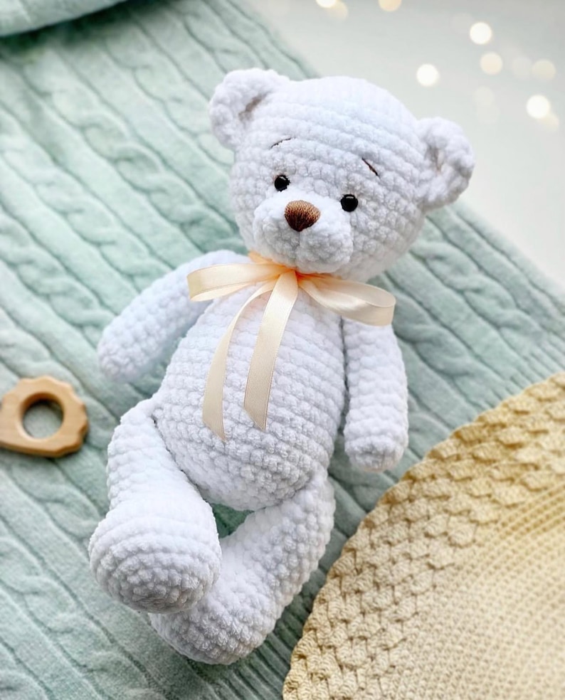 May include: A white crocheted teddy bear with a brown nose and black eyes. The bear is wearing a light beige ribbon around its neck. The bear is sitting on a light blue and beige crocheted blanket.