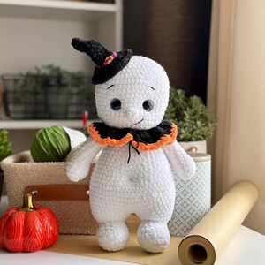 May include: A handmade white crochet ghost doll wearing a black witch hat with orange and pink details. The ghost has a black and orange collar and black button eyes. A red ceramic pumpkin is nearby.