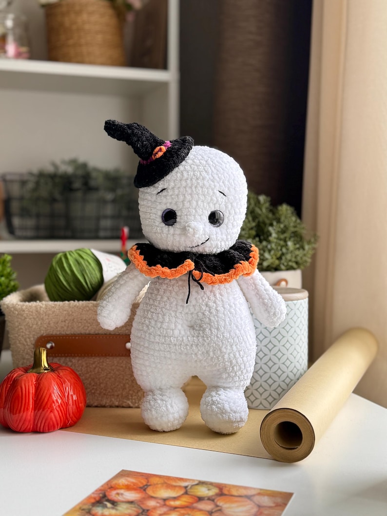 May include: A handmade white crochet ghost doll wearing a black witch hat with orange and pink details. The ghost has a black and orange collar and black button eyes. A red ceramic pumpkin is nearby.