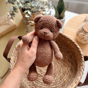 May include: A handmade brown crochet teddy bear with small black eyes and a stitched nose. The bear is held in a woven basket with brown leather handles. The bear is approximately 7 inches tall.